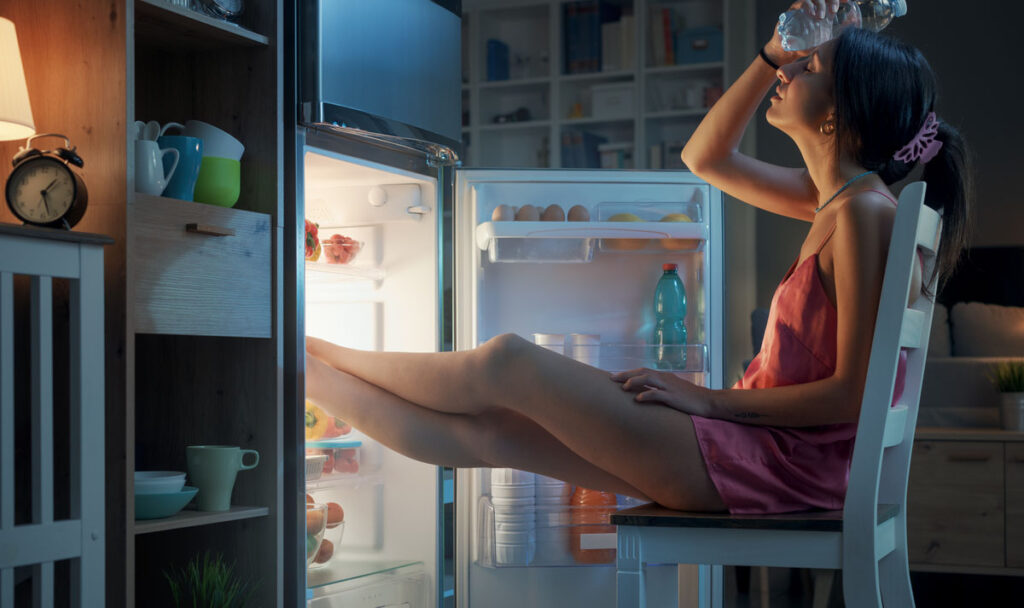 Woman suffering from the heat in summer, she is resting with her feet in the fridge and holding a water bottle